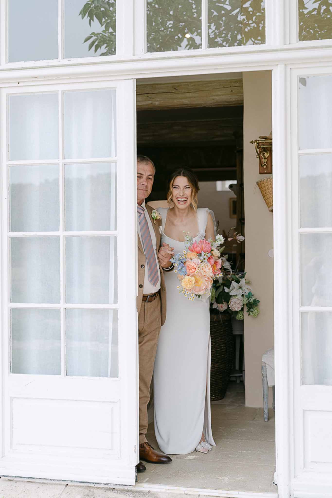 Bride holding coral and peach bouquet in cream column dress, groom in beige suit, framed in glass-paned doorway