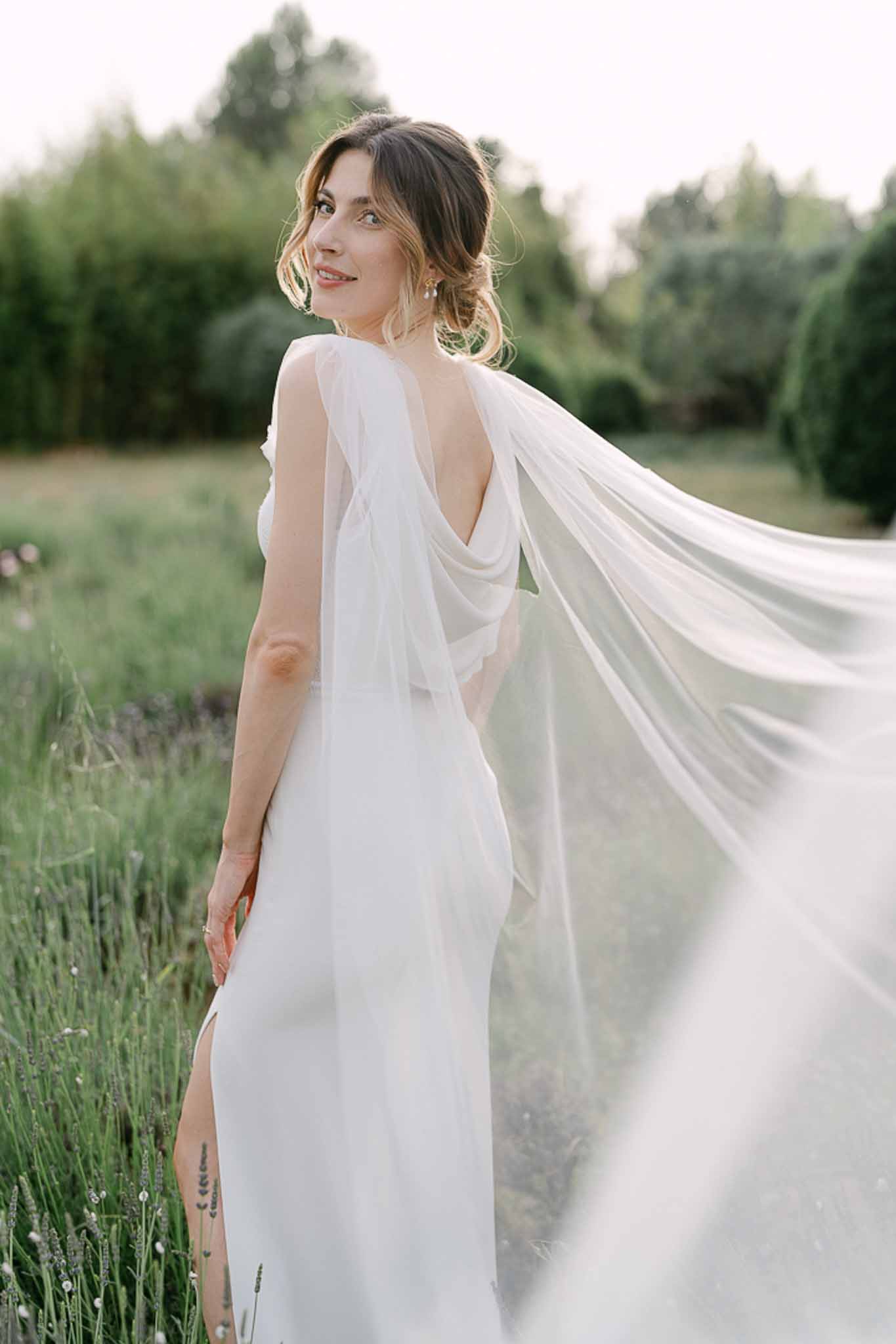 Bridal portrait in lavender field at dusk showing ivory gown with deep V-back and sheer flowing cape