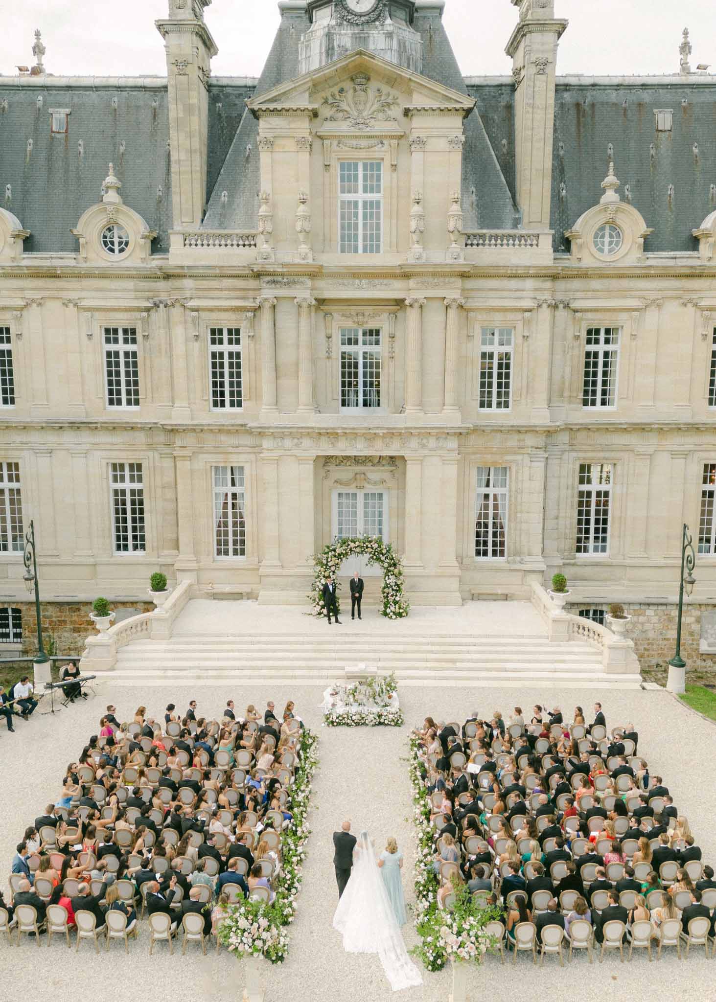 Aerial view of bride walking aisle toward groom under floral arch, 150 guests seated in courtyard of cream stone château