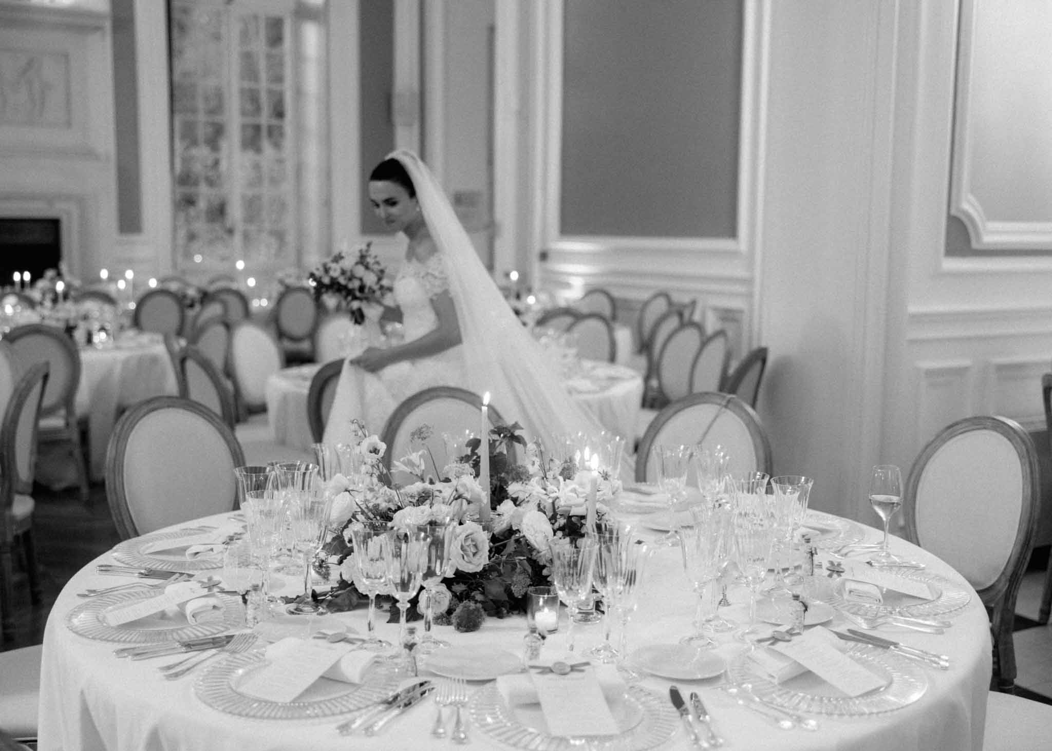 Bride in lace wedding dress seated at a round reception table in a classical ballroom; low floral centerpiece and crystal glassware.