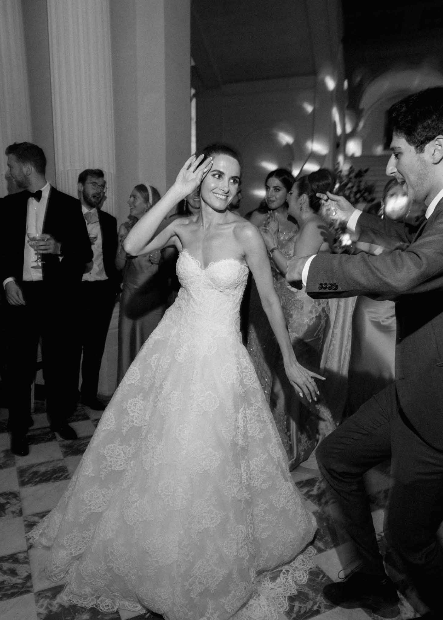 Black and white photo of bride dancing with guests during indoor reception in formal ballroom
