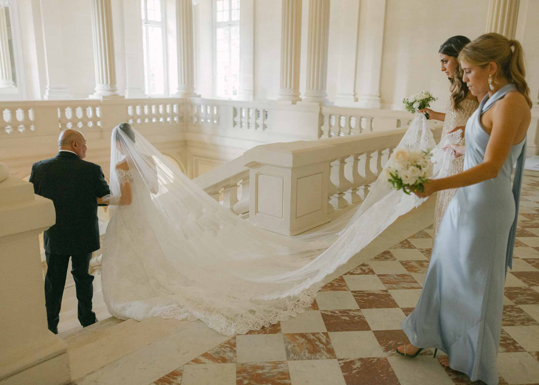 Bride in ivory lace gown with cathedral veil assisted by bridesmaids in a neoclassical hallway with fluted columns.