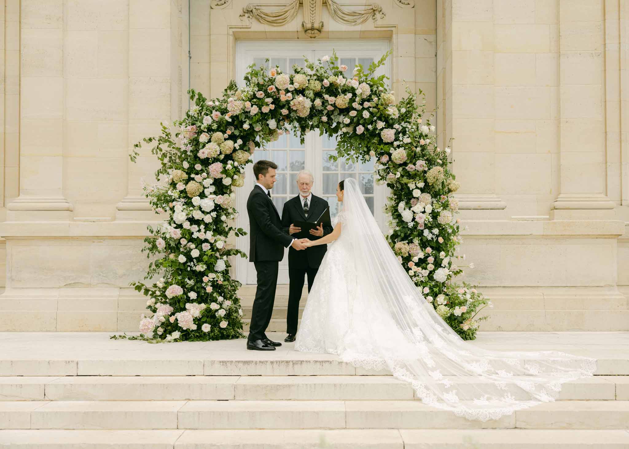Wedding ceremony at stone facade with couple and officiant beneath large cream, blush, and pink floral arch with ivy and roses