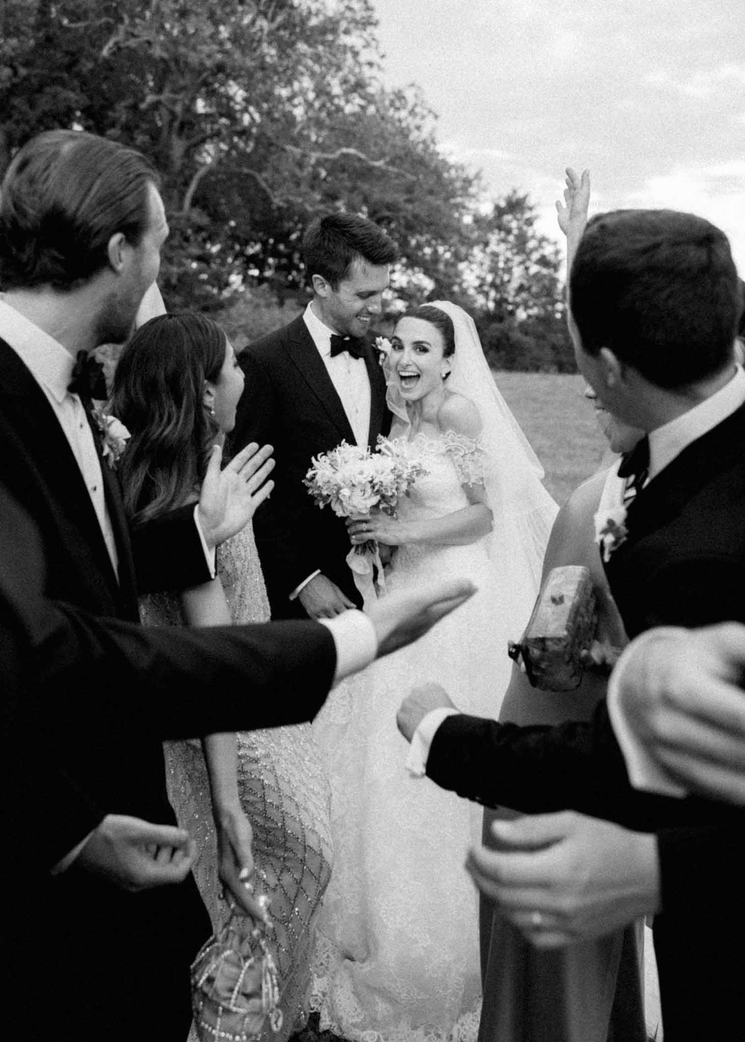 Black-and-white post-ceremony group portrait of bride holding white bouquet surrounded by guests in garden