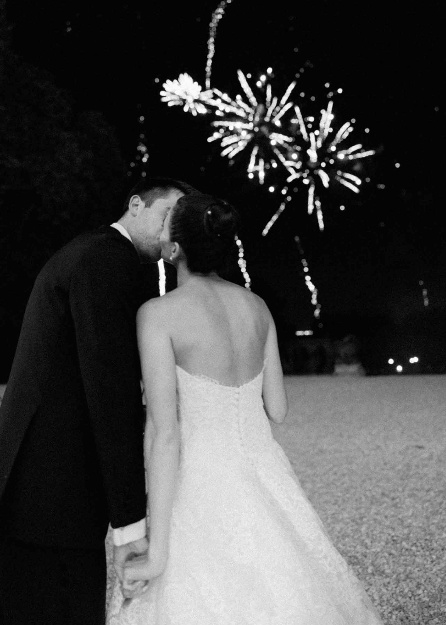 Bride and groom seen from behind watching fireworks burst overhead during evening wedding celebration