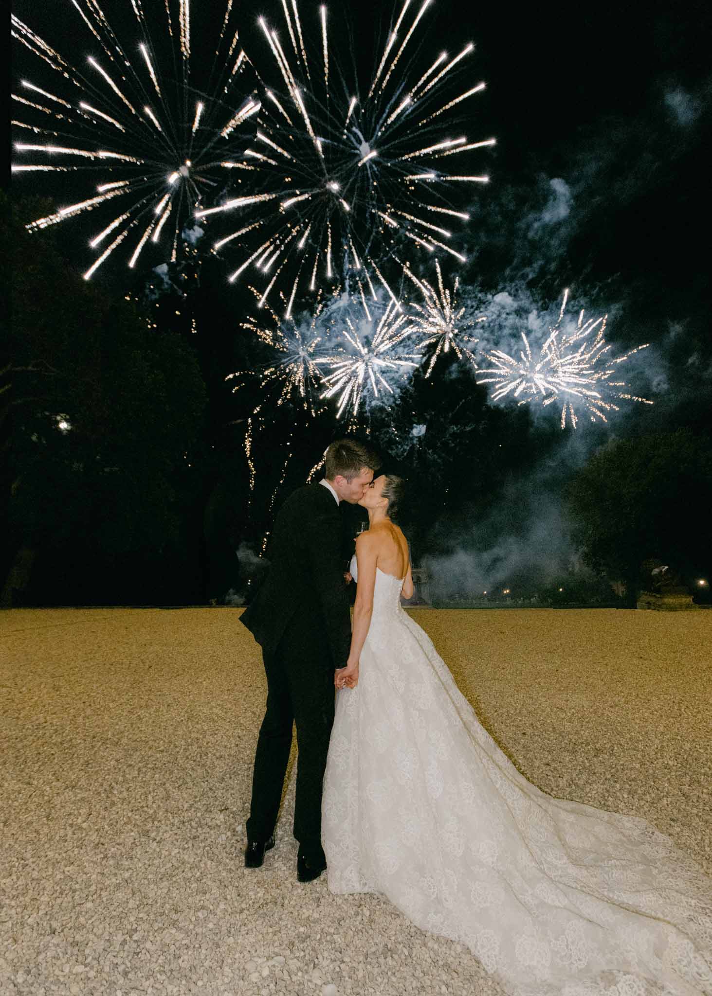 Couple kissing under white and gold fireworks display at night, bride in white strapless gown with long train