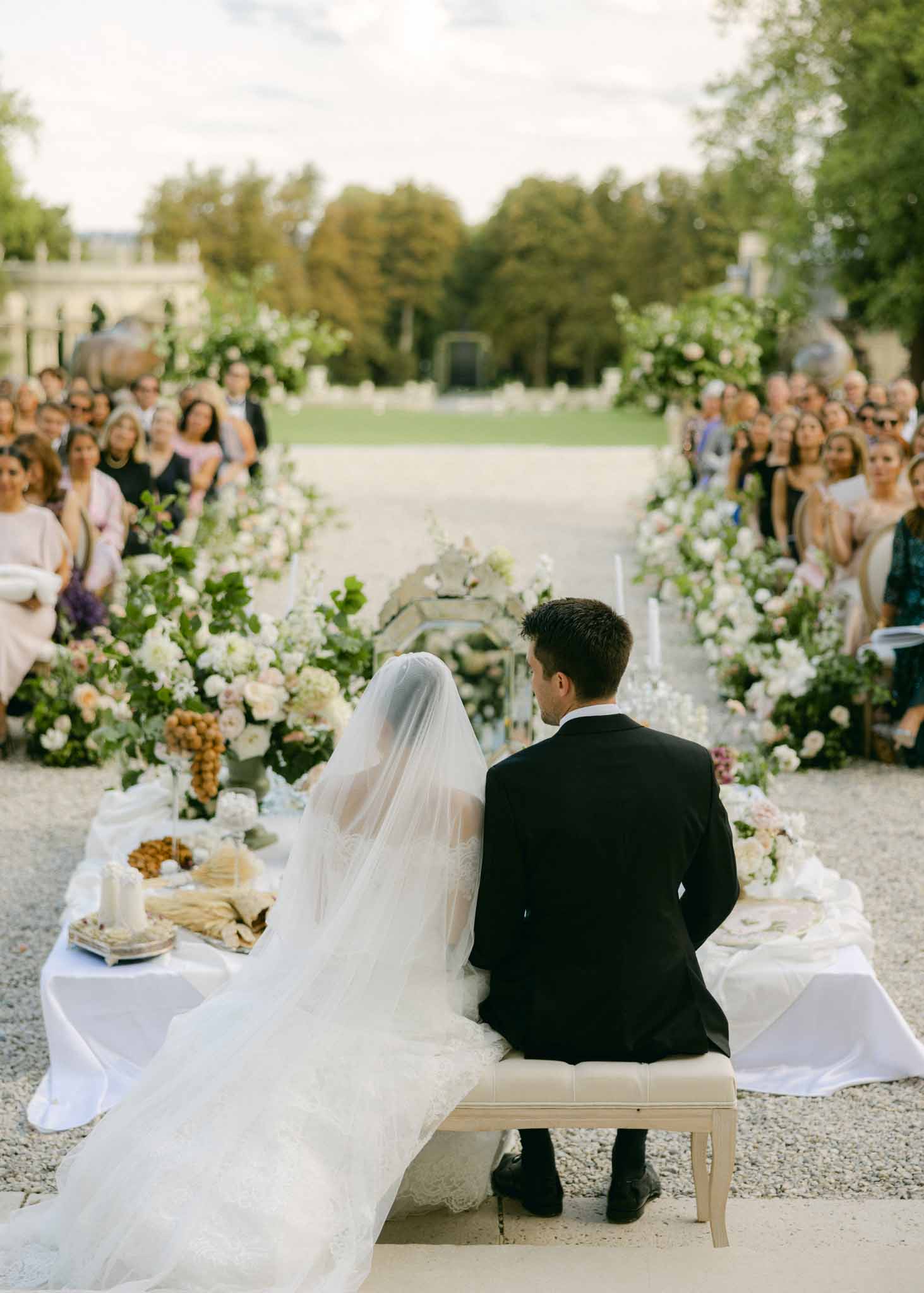Bride and groom on bench facing 60 guests at outdoor ceremony with white and pink florals in gold vessels