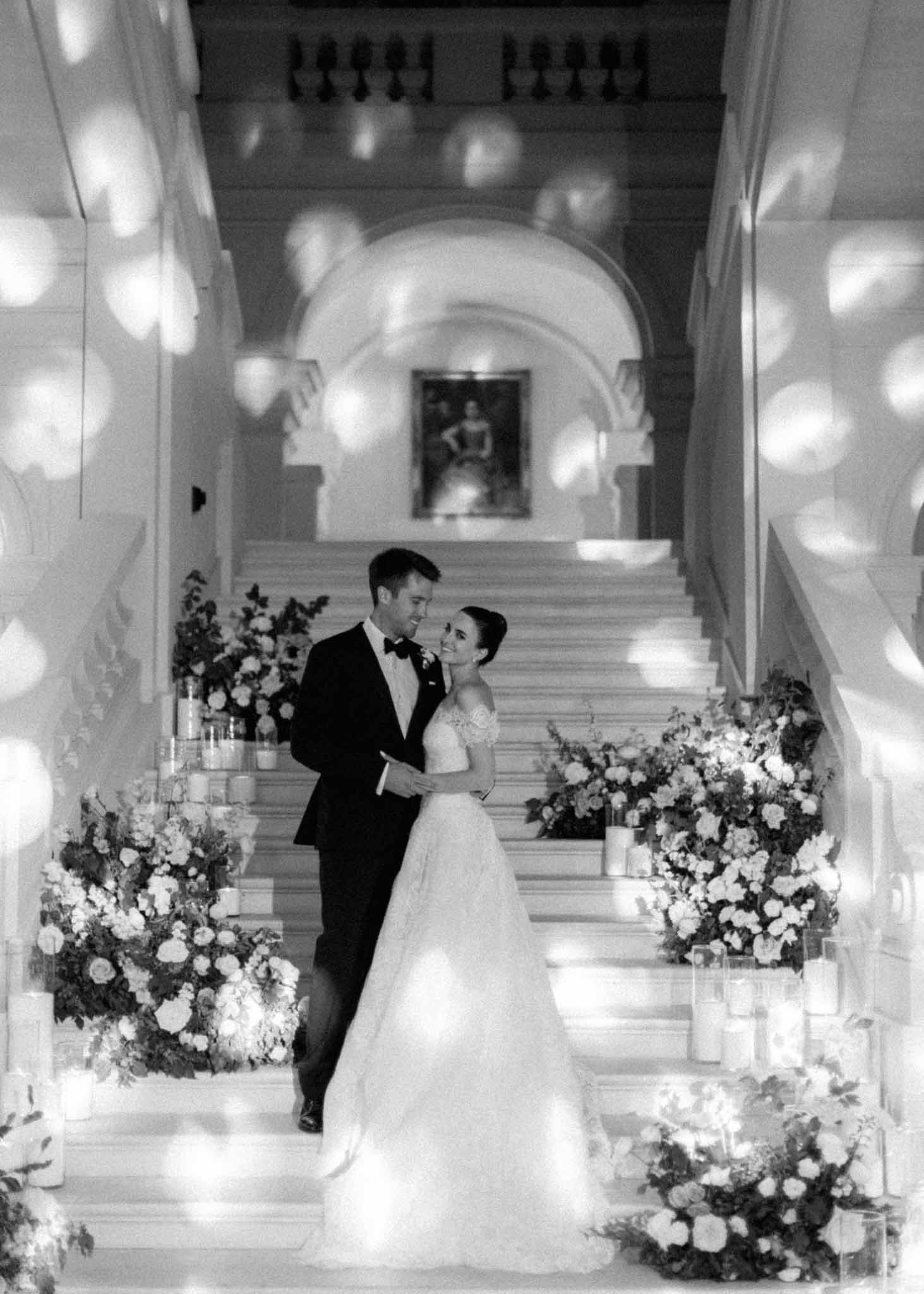 Black and white photo of bride and groom walking through columned hallway with floral arrangements