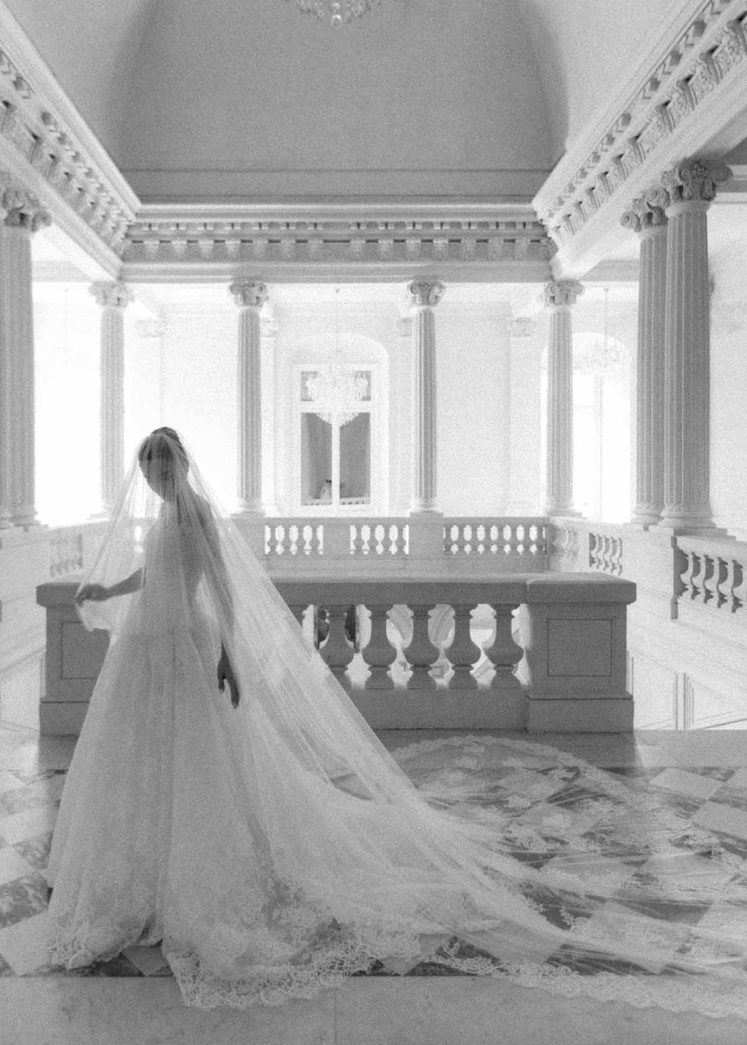 Bride from behind with cathedral-length veil in neoclassical hall with fluted columns and balustrade windows