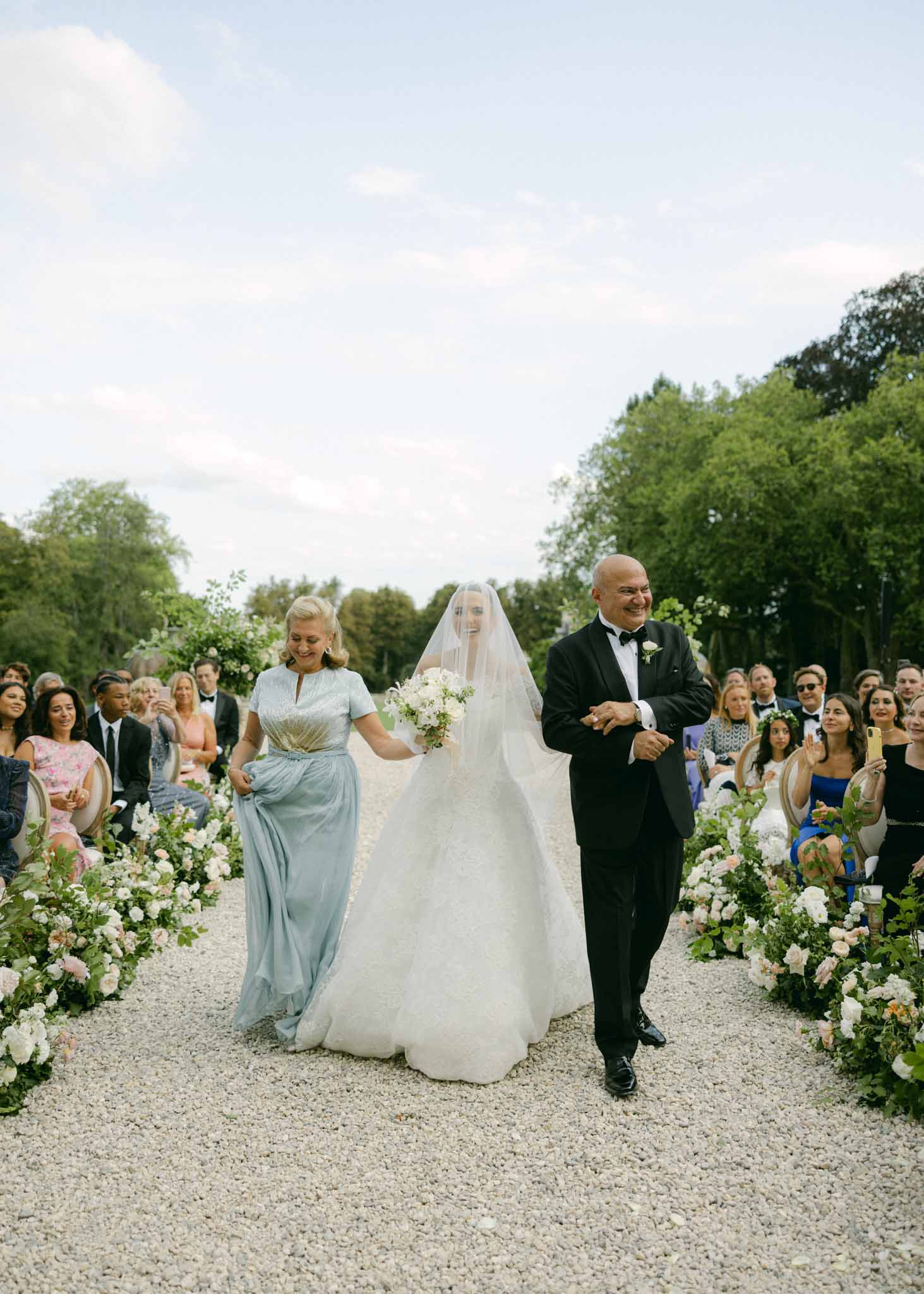 Bride walks down a gravel garden aisle with her parents toward the groom during an outdoor ceremony