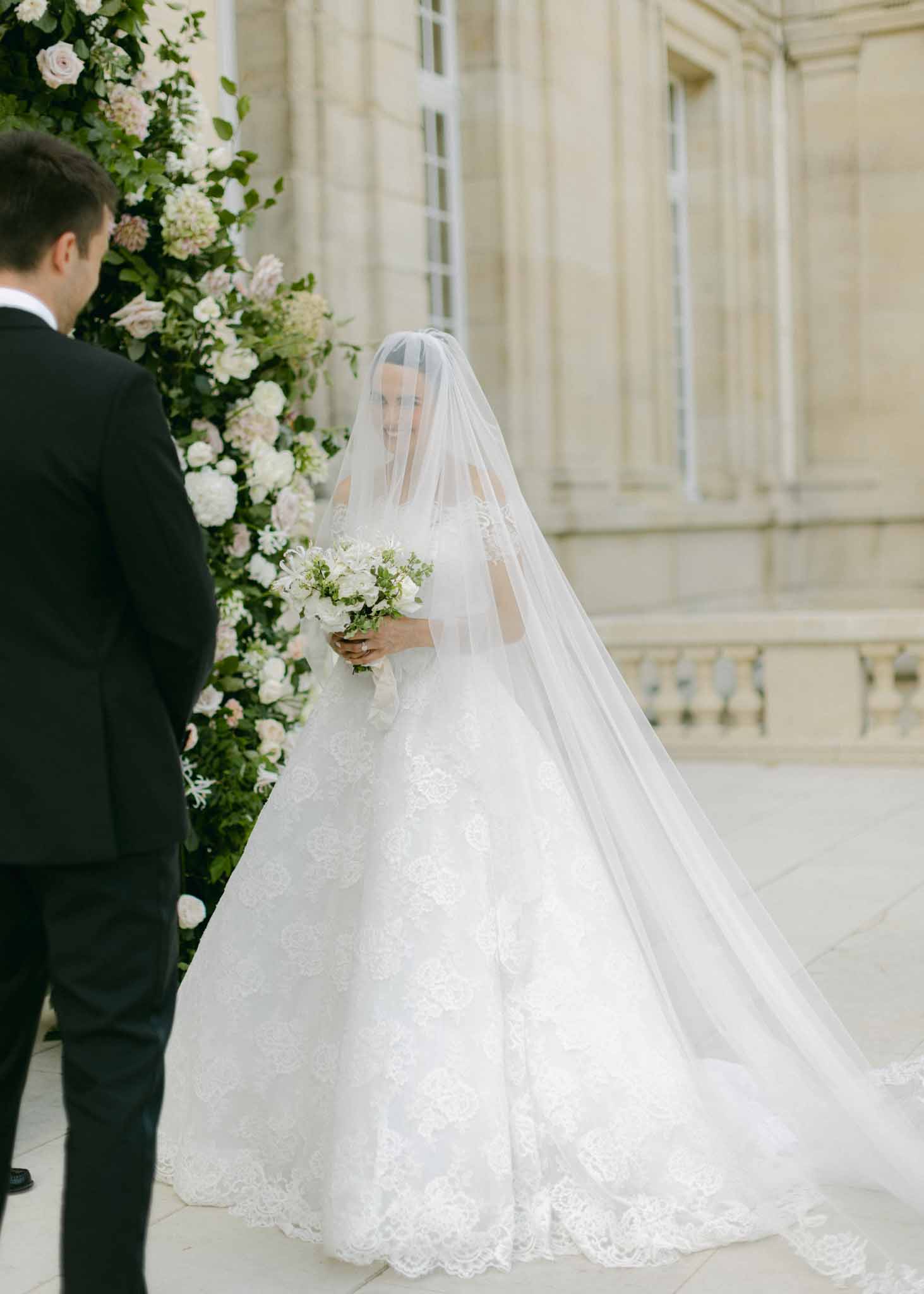 Bride in ivory lace ball gown with veil and groom in black tuxedo at ceremony with tall white and blush floral backdrop
