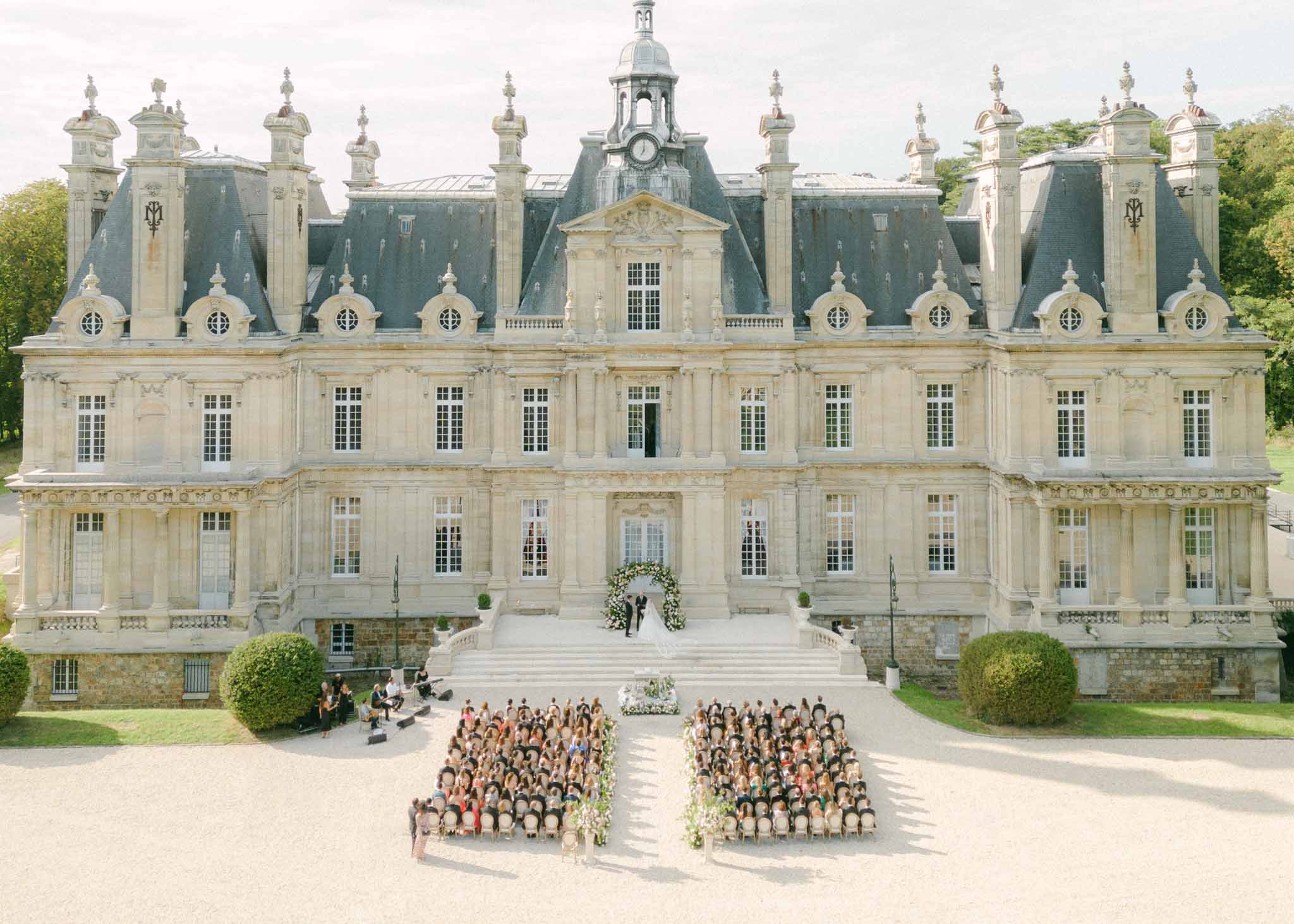 Aerial view of 200 guests at ceremony on chateau courtyard steps, couple under white floral arch at stone staircase