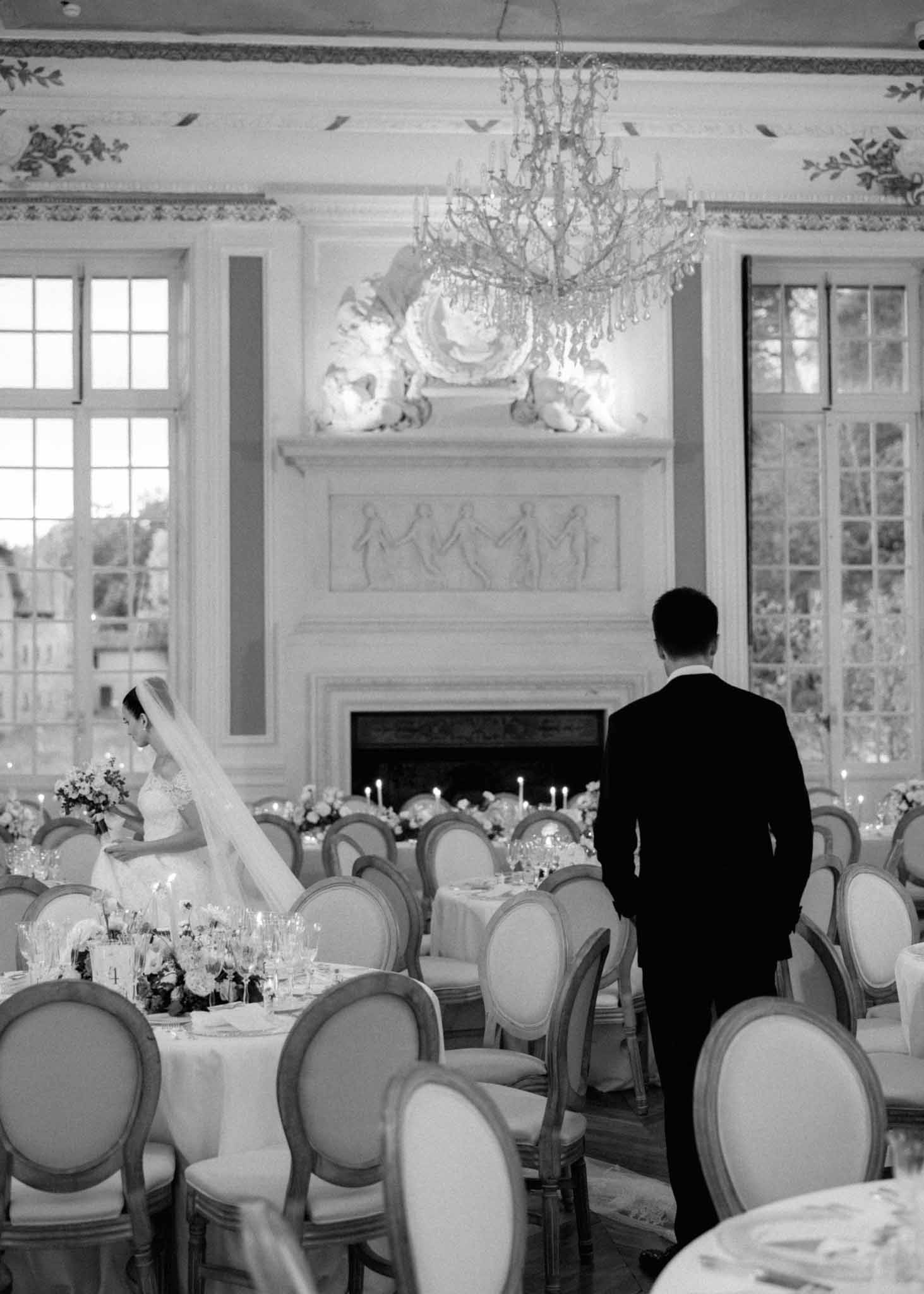 Black-and-white formal reception in neoclassical ballroom with fireplace, chandelier, and seated guests