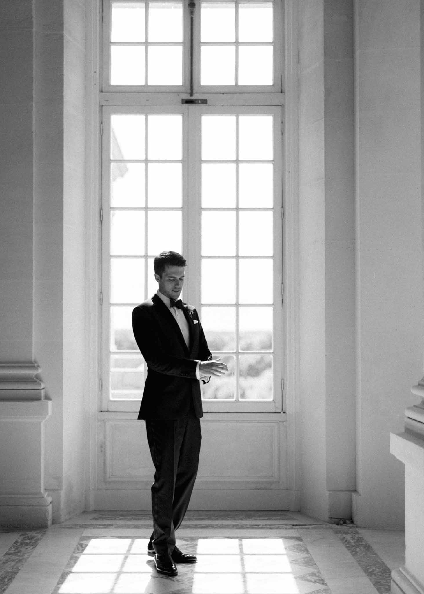 Groom in black tuxedo and bow tie standing in a neoclassical hallway with tall glass doors and geometric tile floor