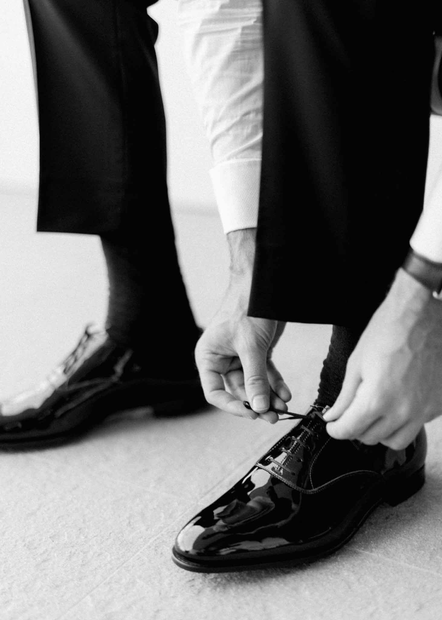 Black-and-white close-up of groom's black oxford shoes being laced during getting ready