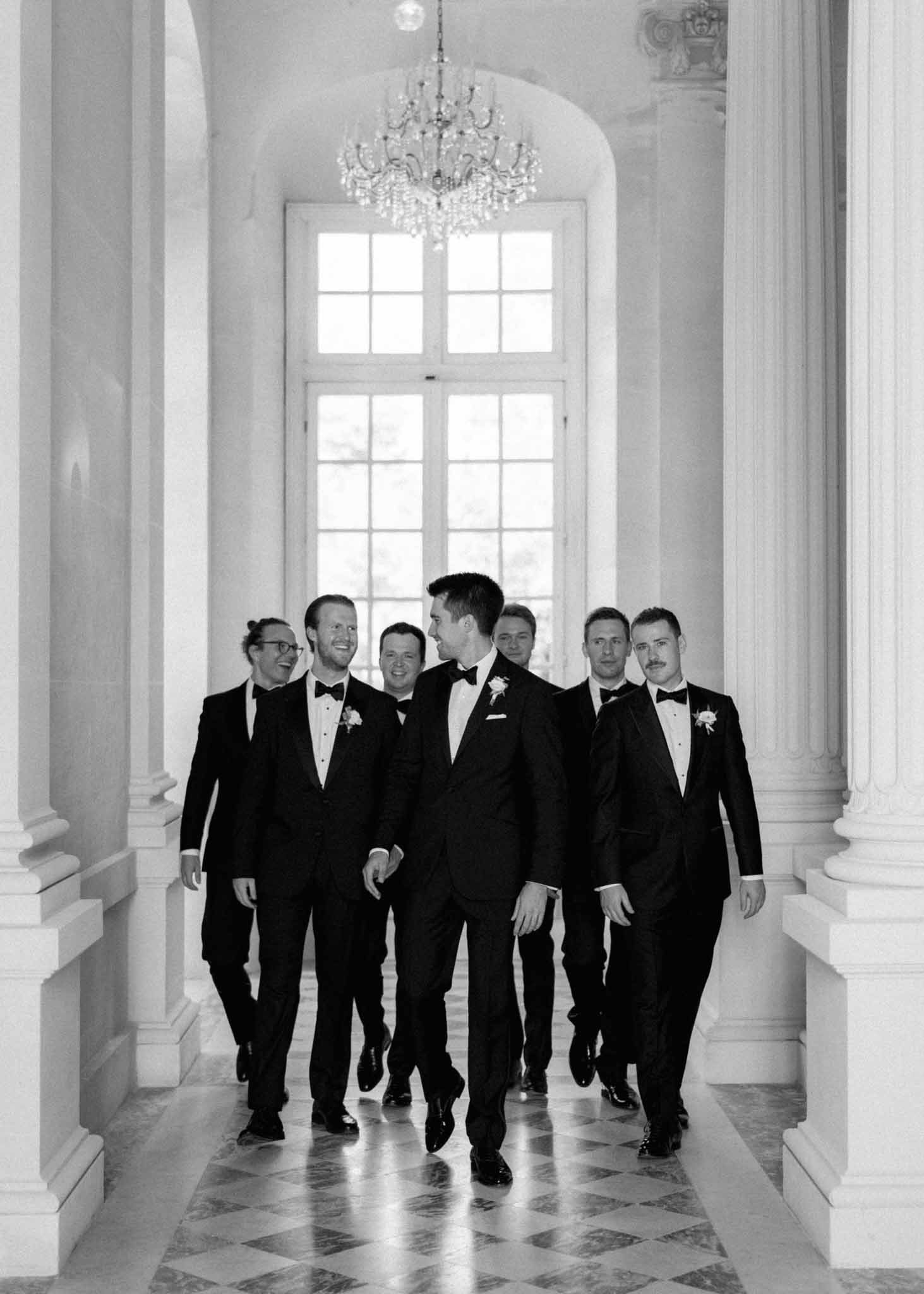 Black-and-white shot of groom and six groomsmen in black tuxedos walking down grand columned château hallway