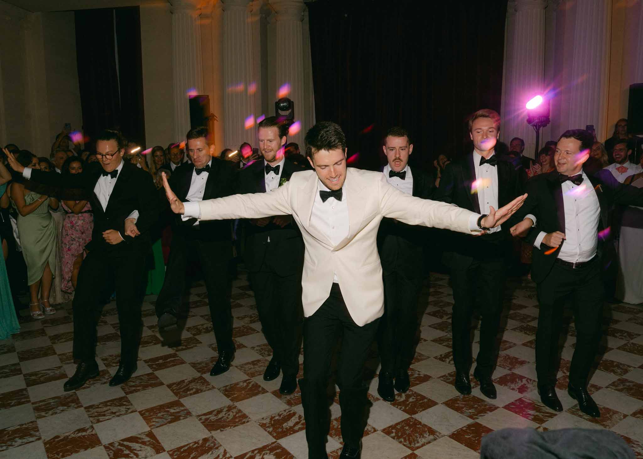 Groom in ivory dinner jacket dancing with arms spread in formal ballroom with pink and purple uplighting