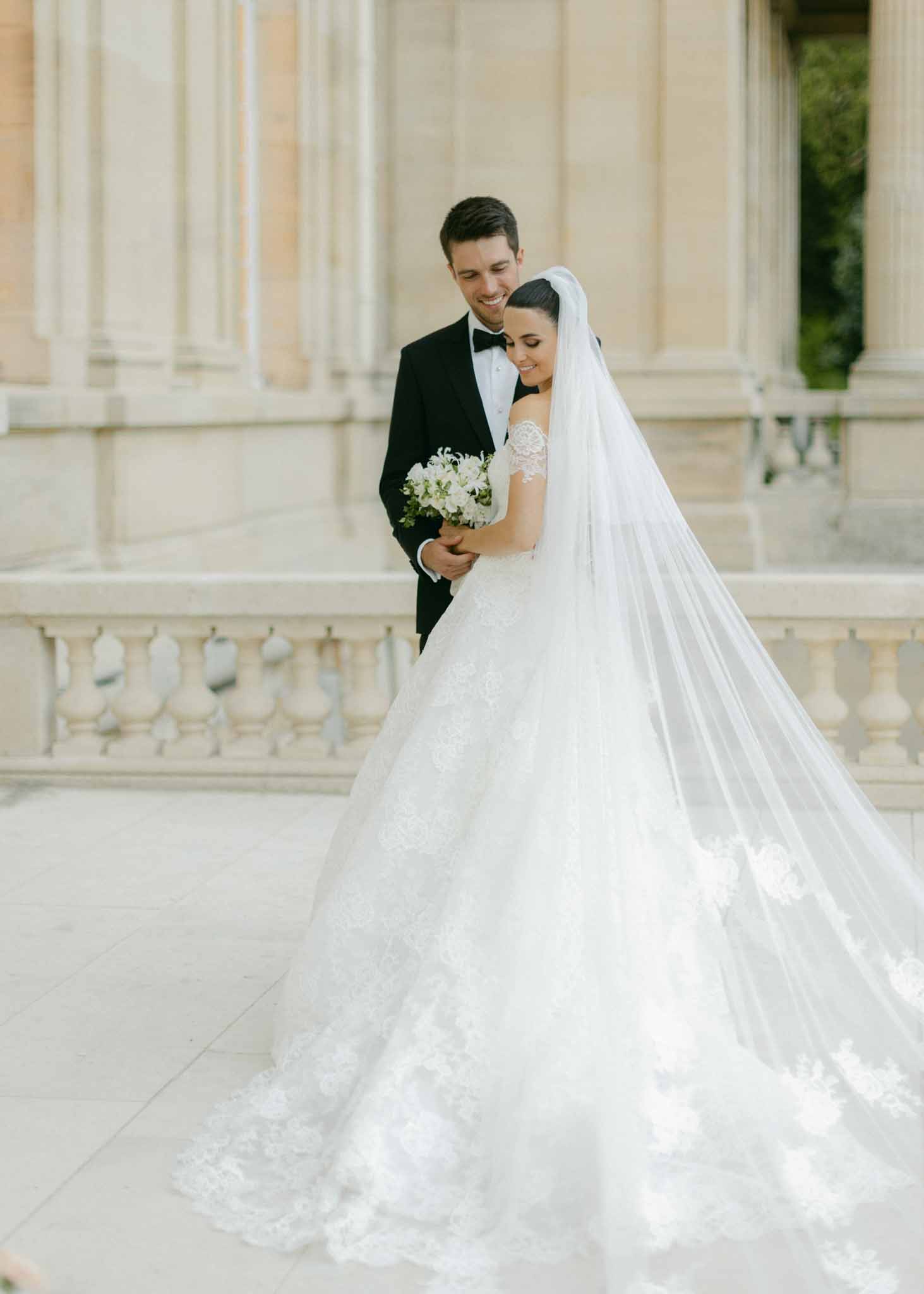Bride in ivory lace gown with full skirt and veil holds white bouquet beside groom on classical stone terrace