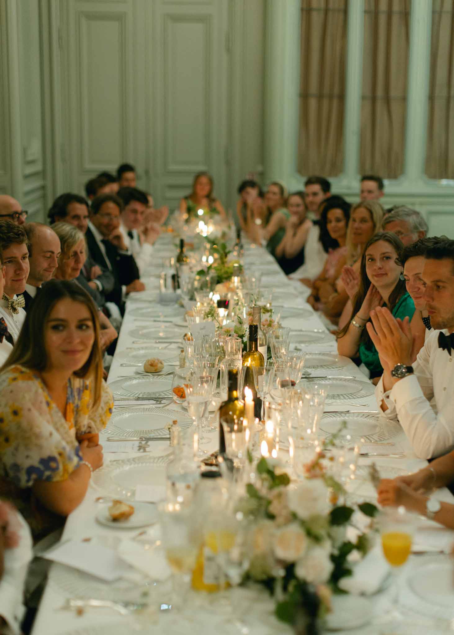Guests at long banquet table in ballroom with pale green paneled walls, white florals and guests in formal attire