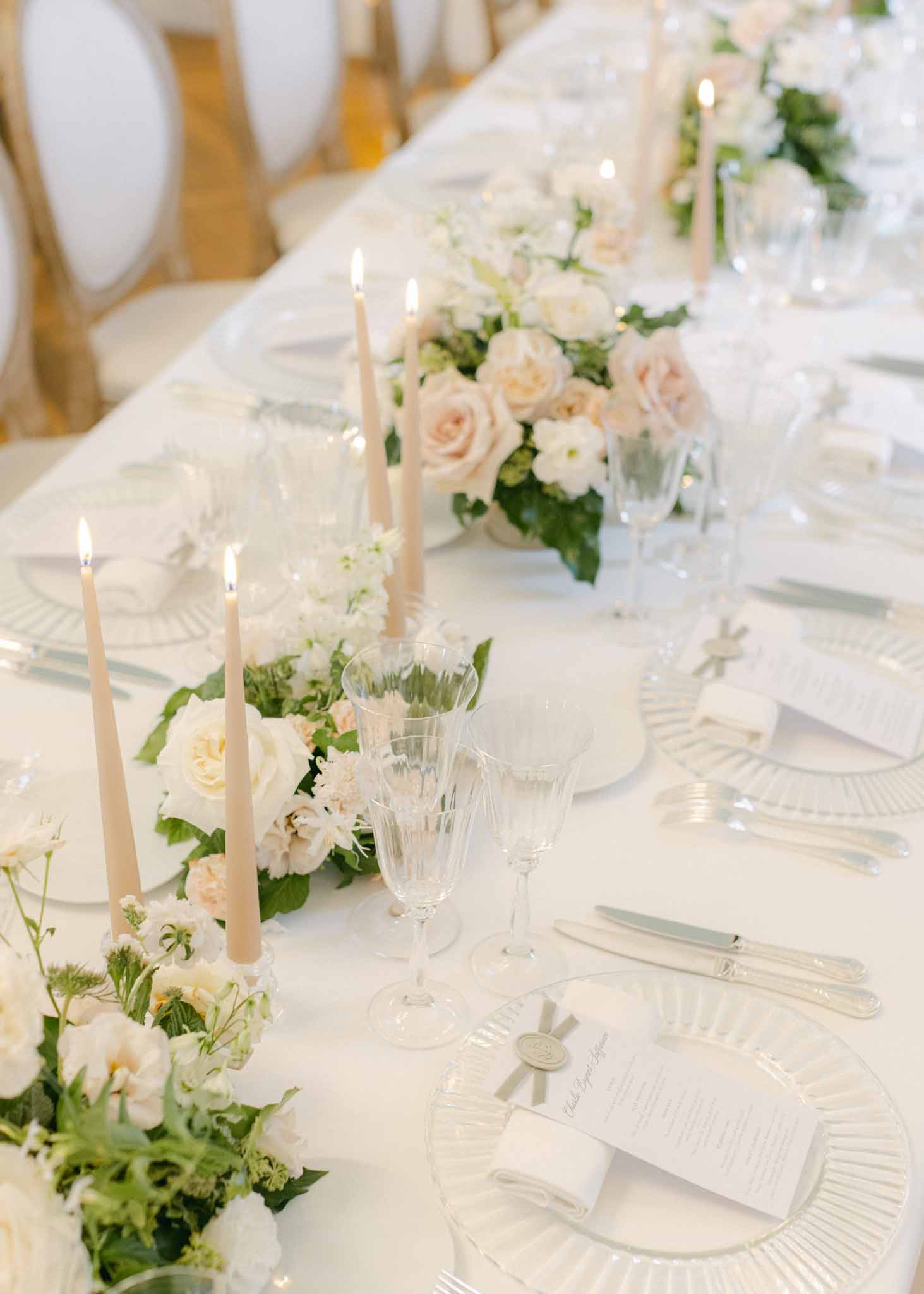Long reception table with white linen and a central garland of cream and blush roses, taper candles and crystal glassware.