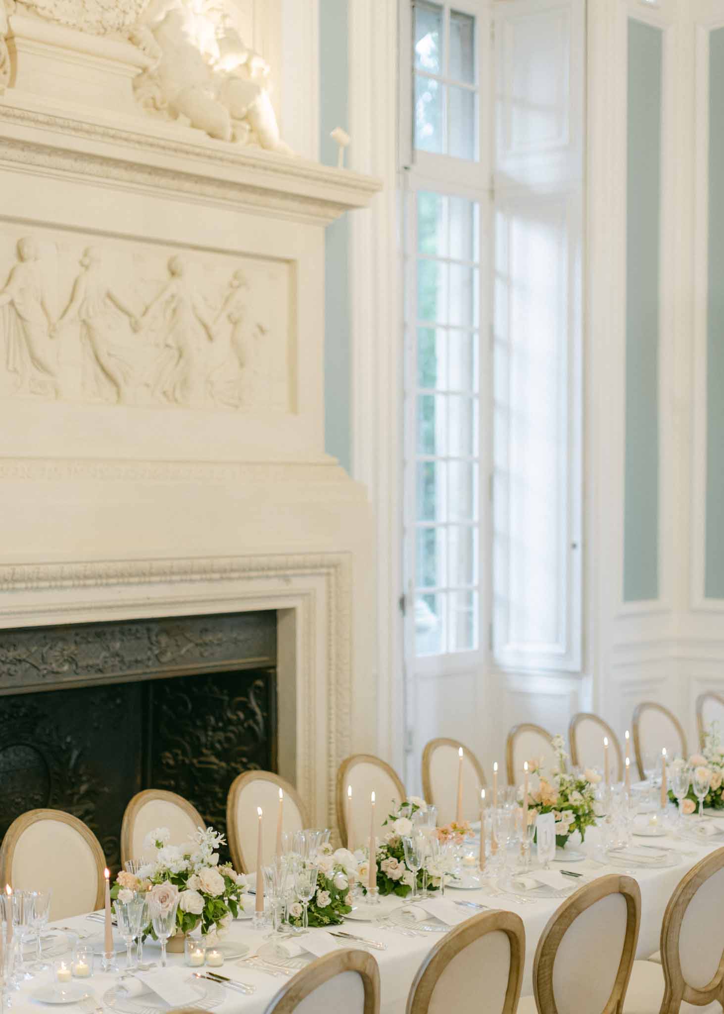 Long banquet table set with white linens and floral centerpieces in a classical ballroom with tall windows