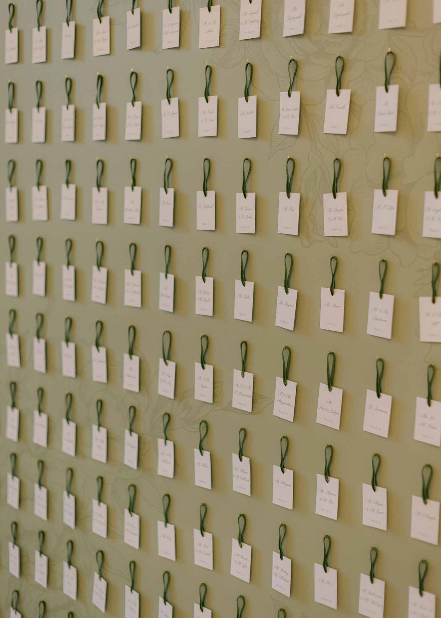 Seating chart with dozens of ivory escort cards suspended from green ribbon loops on a sage green wall