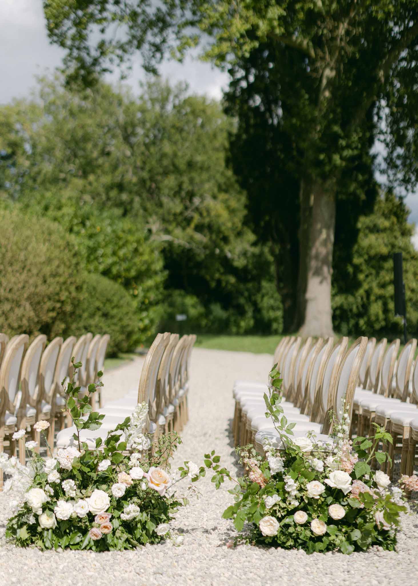 Ceremony aisle with cream wooden chairs, blush rose and baby's breath aisle florals in a tree-lined garden