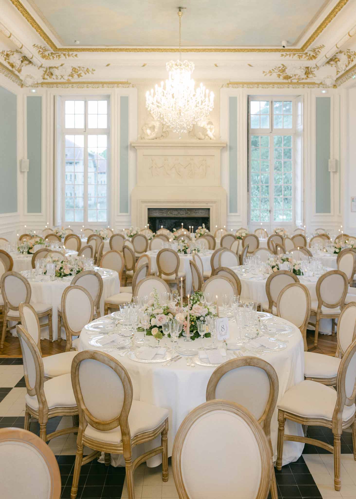 Neoclassical ballroom with crystal chandelier, round tables with white flowers, Louis XVI chairs, and marble fireplace