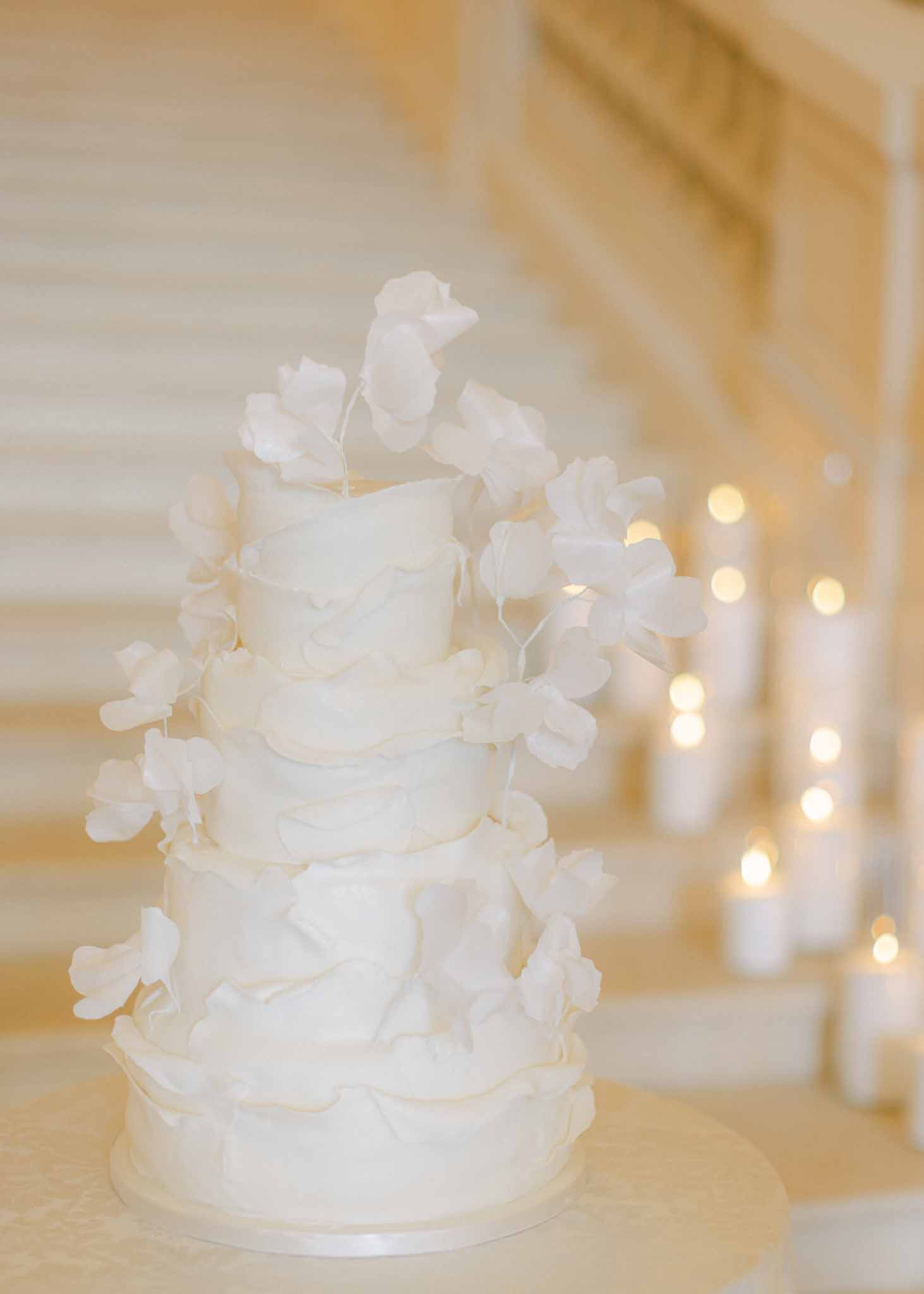 Five-tier white wedding cake adorned with cascading white sweet pea flowers and candlelight