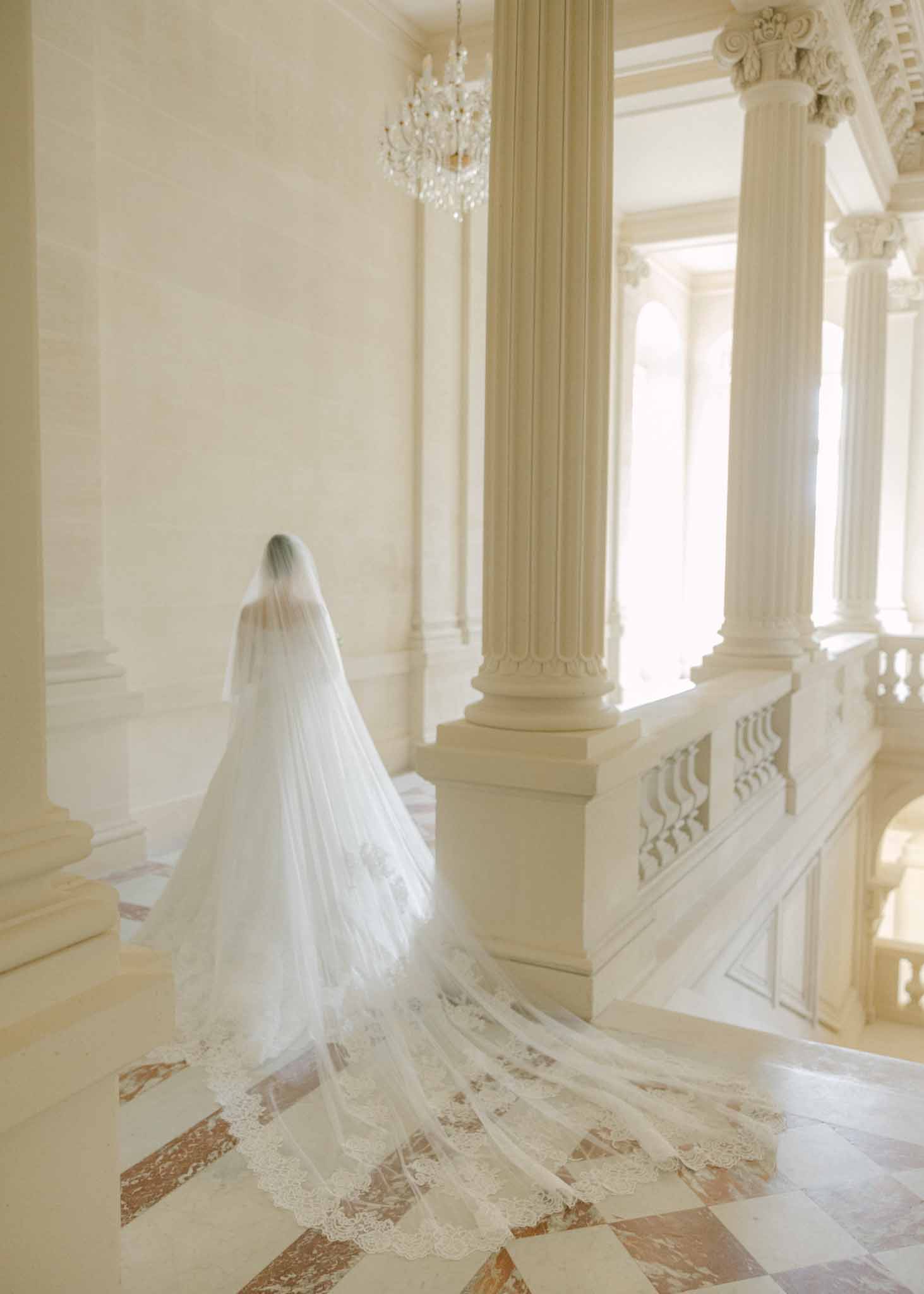 Bride from behind in ivory lace gown with long tulle veil in a neoclassical hallway with columns and crystal chandelier.
