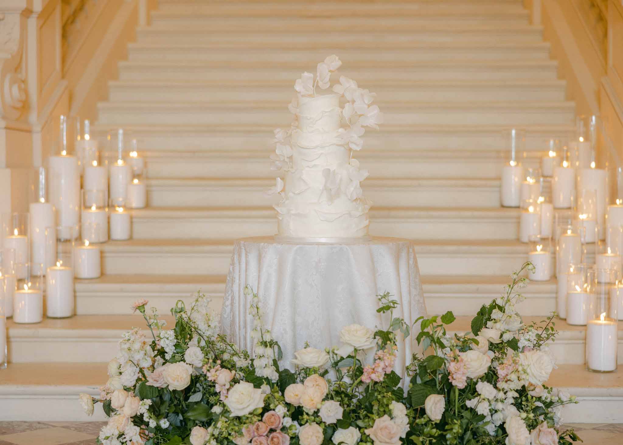 Three-tiered white wedding cake surrounded by ivory and blush roses with pillar candles in glass hurricanes