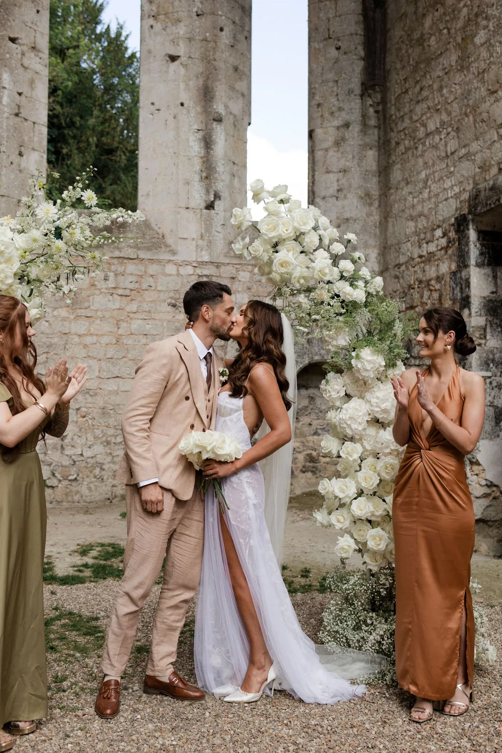 Bride and groom kissing in stone courtyard with bridesmaids in earth tones and ivory rose floral arch