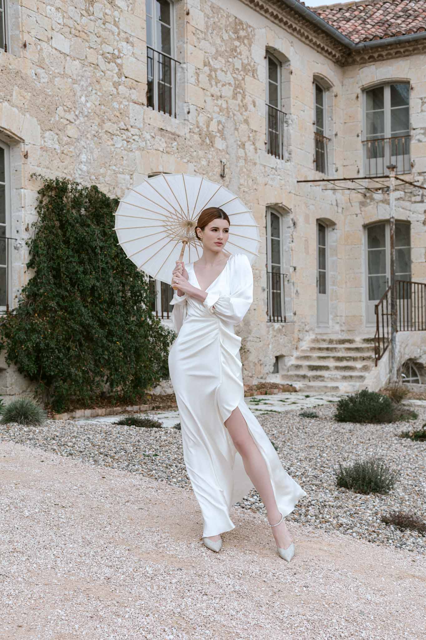 Full-length bridal portrait with cream parasol in historic stone chateau courtyard with arched windows
