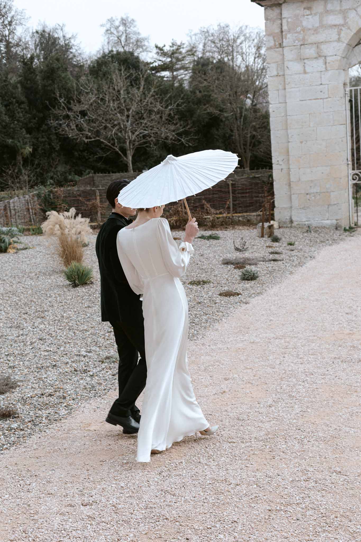 Bride in ivory silk gown with umbrella and groom walking through gravel courtyard in winter