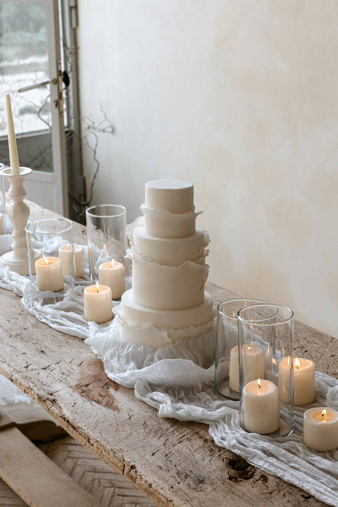 Five-tiered ivory scalloped wedding cake with ruffles on wooden windowsill surrounded by cream candles and ivory tulle