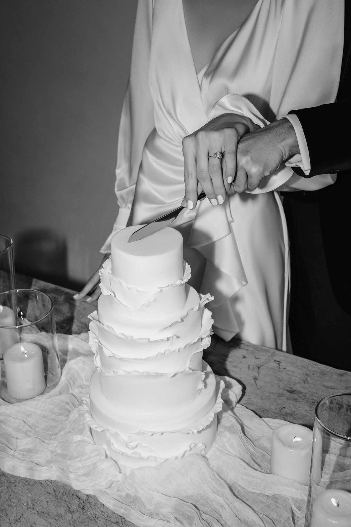 Black-and-white photo of couple cutting five-tier white wedding cake with ruffled frosting and candlelight
