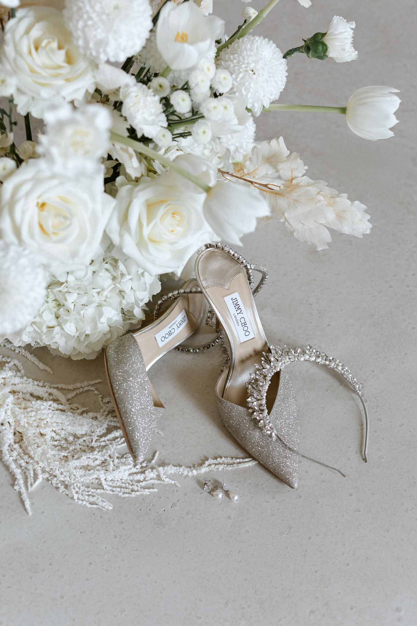 Flat lay of gold glitter bridal heels with rhinestone straps beside white rose and dahlia bouquet