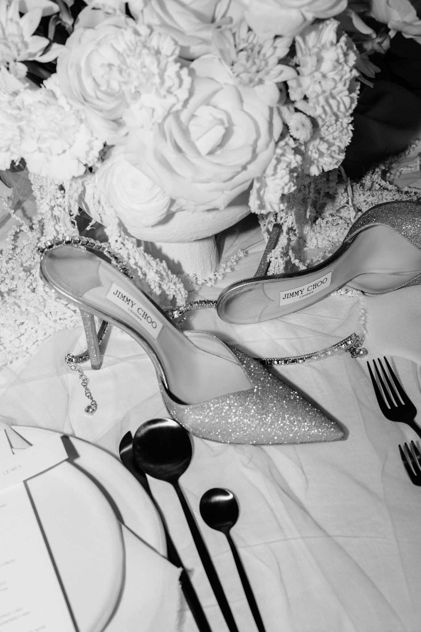 Silver glitter heels with rhinestone straps beside white tiered wedding cake with roses, black-and-white flat lay