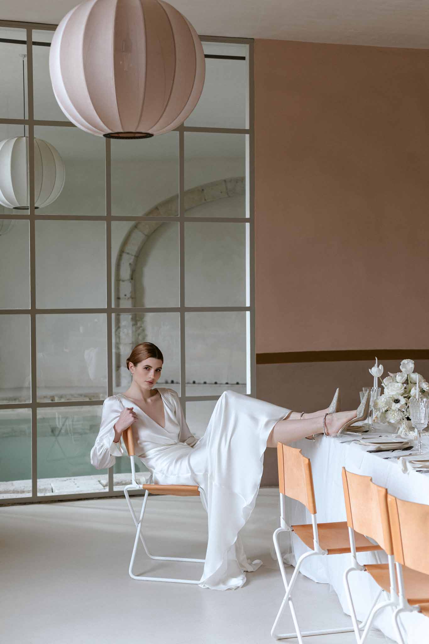Bride in cream silk wrap dress seated at minimalist reception table with paper lanterns and gridded windows at indoor venue