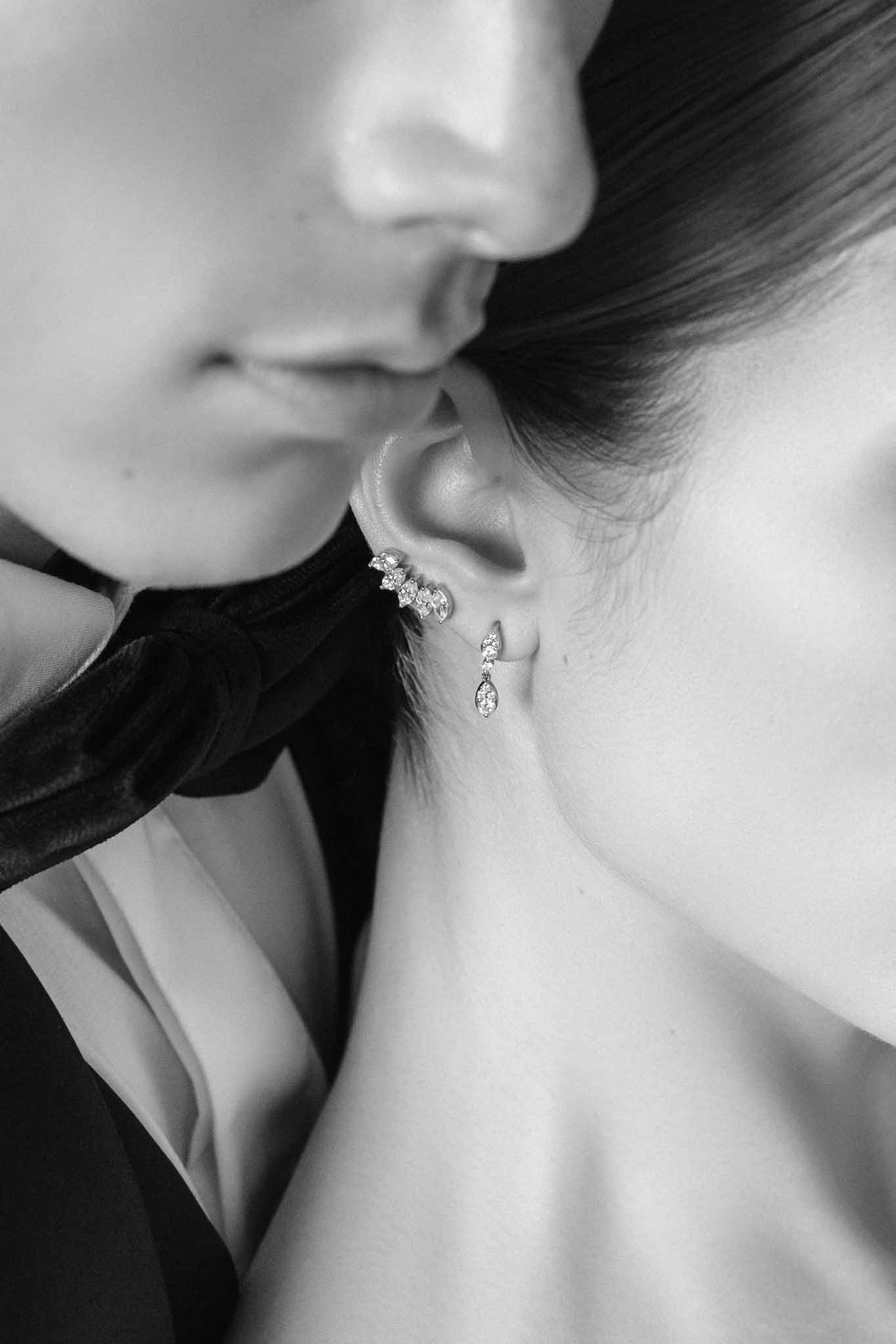 Close-up of bride ear with marquise diamond earrings as second person adjusts jewellery, black-and-white