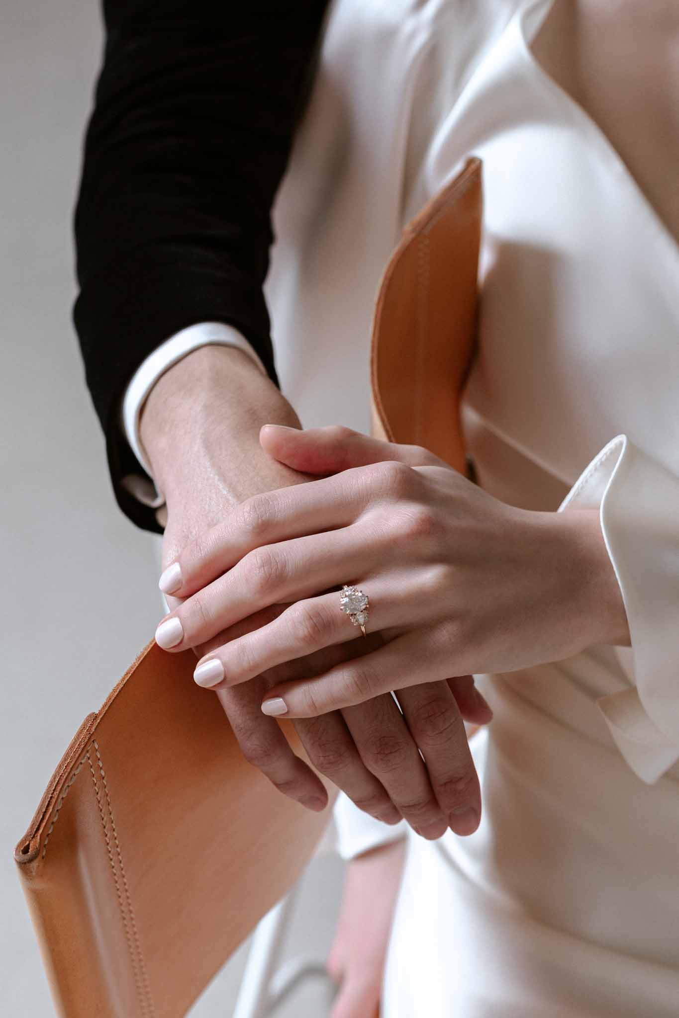Close-up of couple's hands showing wedding rings with bride's halo diamond engagement ring and tan clutch