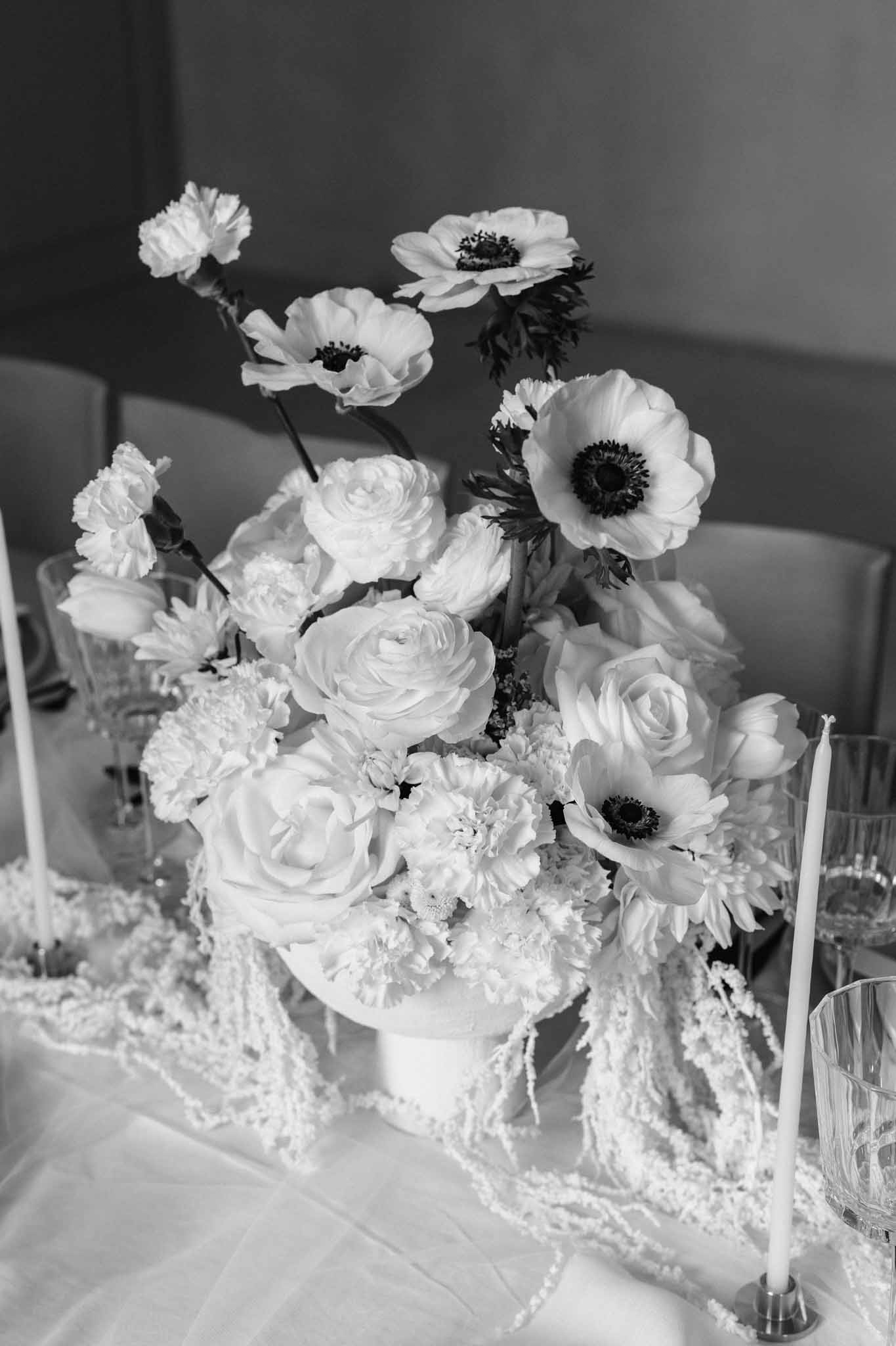 Black and white close-up of white roses, carnations and anemones in a white vessel with beaded garland on a reception table.