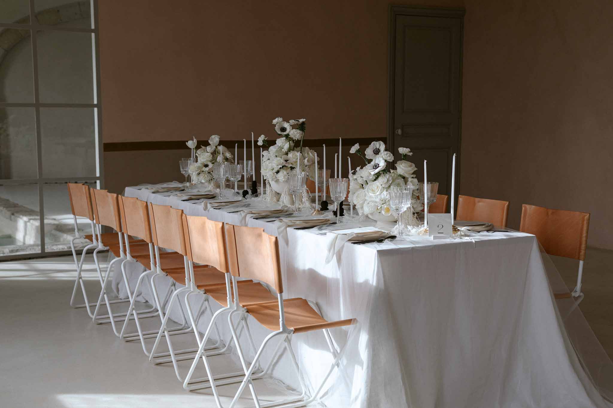 Long reception table with white linens, apricot runner, taper candles, and white floral centerpieces in a minimalist room