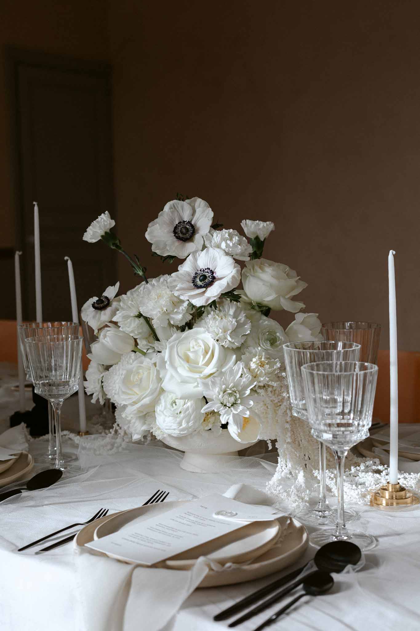 Winter wedding place setting with white ceramic compote, ivory roses, black flatware, and brass candleholders