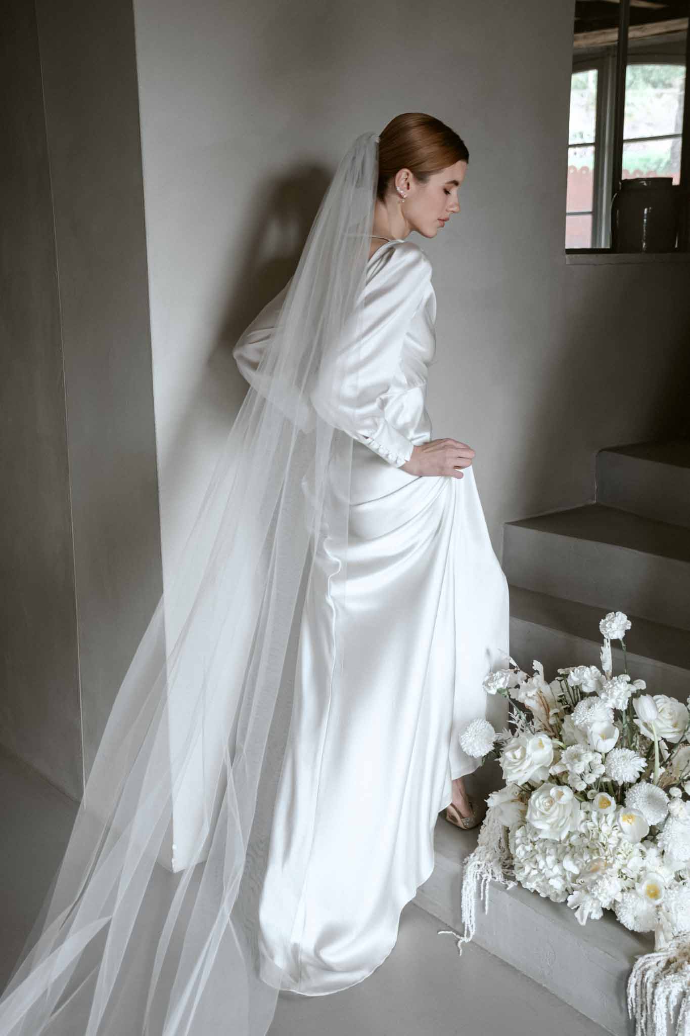 Bride in profile wearing ivory satin gown with long tulle veil and white rose bouquet