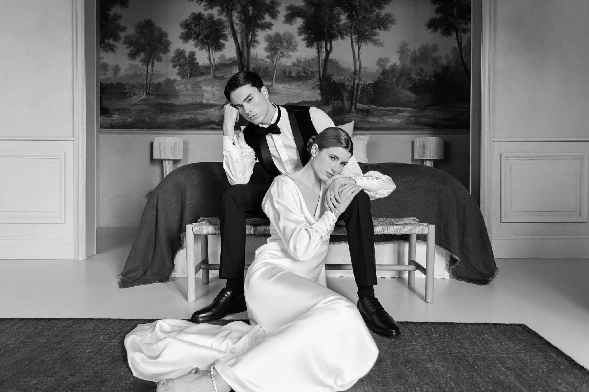 Black and white editorial portrait of couple on bed, bride in white dress and boots, groom in tuxedo