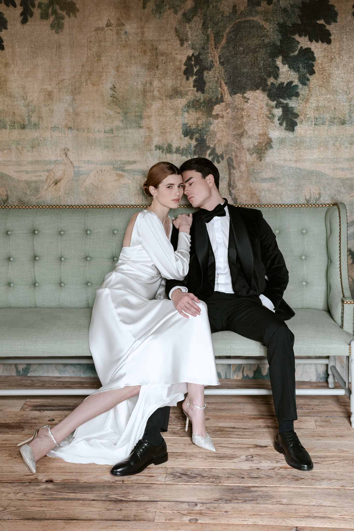 Bride in ivory bias-cut silk dress and groom in black tuxedo seated on sage green sofa with botanical mural behind them