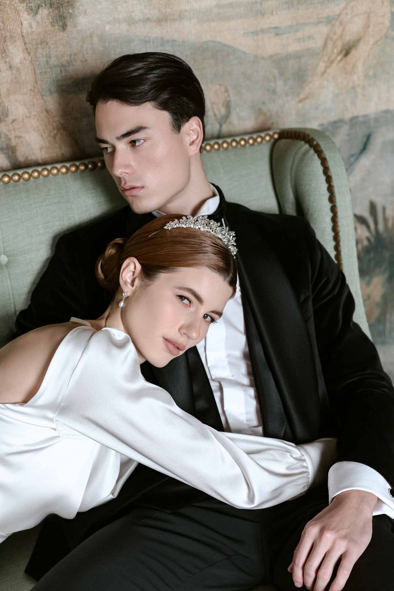 Bride in ivory silk gown with crystal tiara and groom in black tuxedo seated on sage green chair