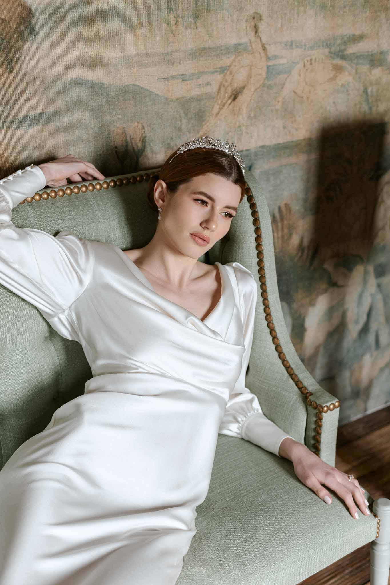 Bride in ivory satin dress with crystal tiara seated on sage green chair by classical mural