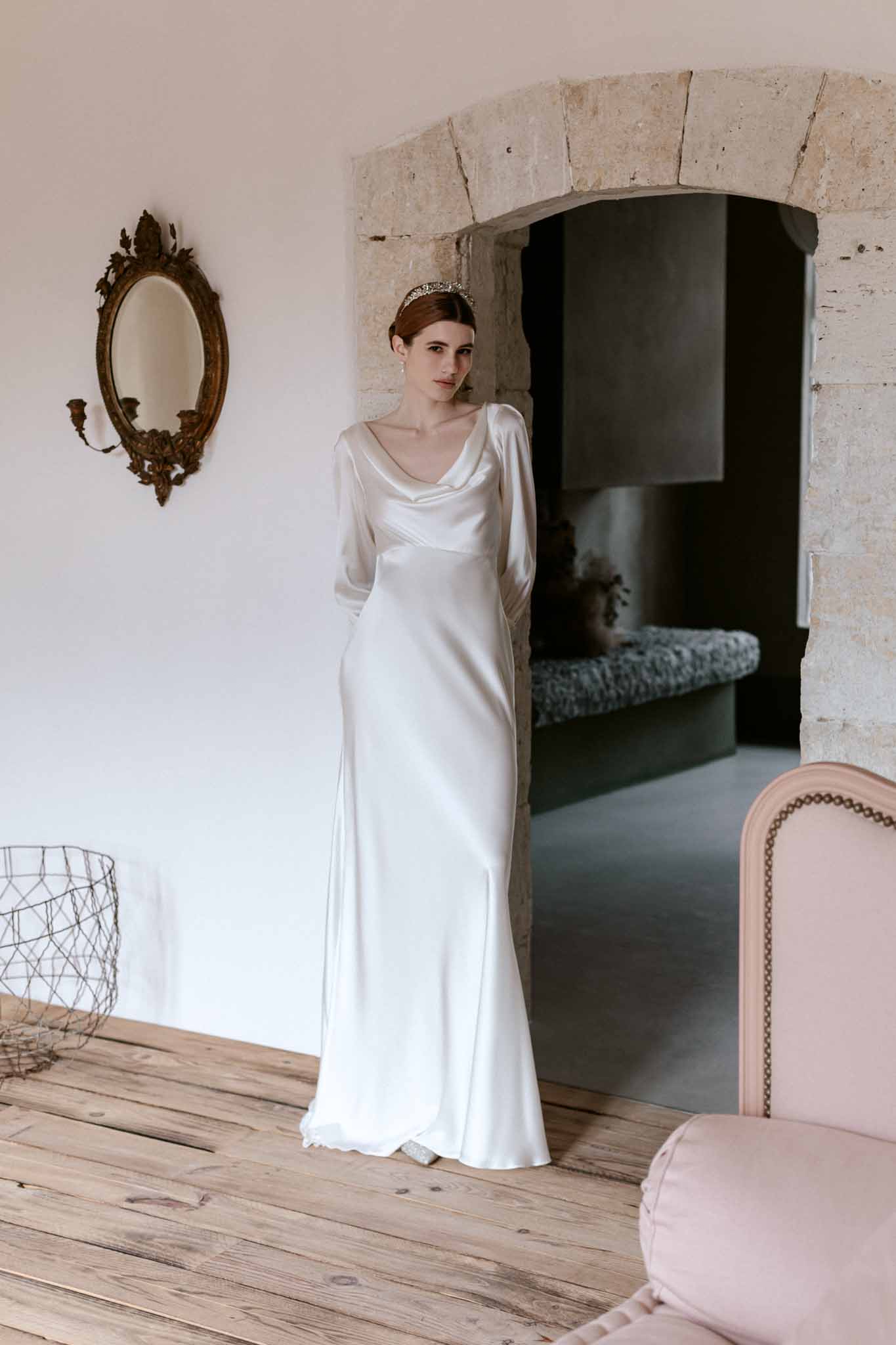 Bride in long-sleeved ivory silk gown with gold headpiece standing in a stone-walled room with gilt mirror